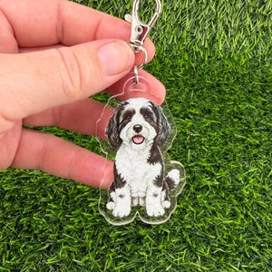 May include: A clear acrylic keychain featuring a black and white dog illustration. The dog is sitting with its tongue out. The keychain has a silver clasp for attaching to keys or bags. The background is green.