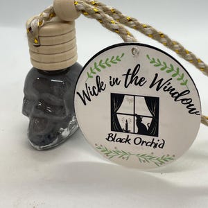 Puede incluir: Un ambientador para coche con forma de calavera, tapa de madera y cordón trenzado. La etiqueta dice "Wick in the Window" y "Black Orchid" con un dibujo de un gato en una ventana.