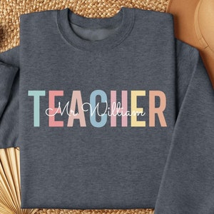 May include: A dark blue sweatshirt with the word "TEACHER" in rainbow colors and the name "Mr. William" written in cursive below.