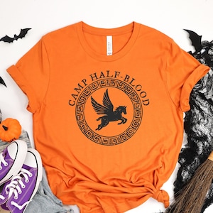May include: Orange t-shirt with the text "Camp Half-Blood" in a black circle with a winged horse inside. The shirt is on a white background with Halloween decorations.