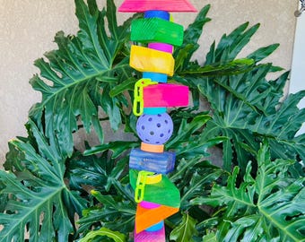 May include: A colorful bird toy with a pink pyramid top, green, yellow, and pink blocks, and a purple ball. The toy is suspended from a chain and is surrounded by green leafy plants. The text "4a" is in the upper left corner.