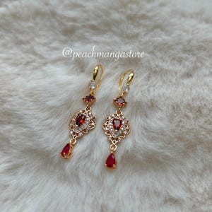 May include: A pair of gold-colored drop earrings with red and white gemstones. The earrings have a decorative design with a teardrop-shaped red gem at the bottom. The text "@peachmangastore" is visible.