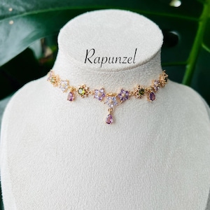 May include: A gold-toned choker necklace with floral and teardrop accents. The necklace features purple, green, and clear gemstones. The word "Rapunzel" is written in cursive script above the necklace.