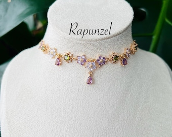 May include: A gold-toned choker necklace with floral and teardrop accents. The necklace features purple, green, and clear gemstones. The word "Rapunzel" is written in cursive script above the necklace.