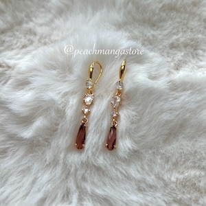 May include: A pair of gold-colored dangle earrings with clear and maroon-colored gemstones. The earrings have a lever-back closure and are displayed on a white, fluffy surface. The text "@peachmangastore" is visible.