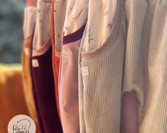 Puede incluir: Una fila de pantalones de pana en varios colores, incluyendo mostaza, burdeos, óxido, malva y verde salvia. Cada par cuenta con una cintura ancha y bolsillos forrados con una tela crema estampada con un delicado patrón botánico marrón. Una pequeña etiqueta con un icono de wombat y el texto Cozy Wombat es visible en los bolsillos.