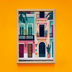 May include: A framed illustration of colourful buildings with balconies and doors. The buildings are painted in shades of pink, blue, and yellow, with green plants and trees in the foreground. The illustration is framed in a light wood frame.