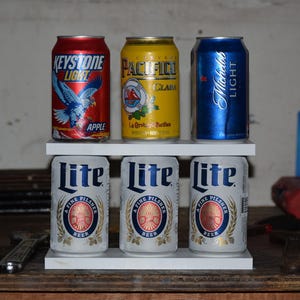 May include: A two-tiered white shelf holding six aluminum beverage cans. The top row features a red Keystone Light Apple can, a yellow Pacifico Clara can, and a blue Michelob Light can. The bottom row contains three white Miller Lite cans.