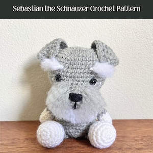 Könnte beinhalten: Ein gehäkeltes graues und weißes Miniatur-Schnauzer-Spielzeug. Der Hund sitzt mit gekreuzten Pfoten und hat eine schwarze Nase und Augen.