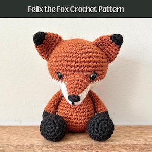 May include: A crocheted fox amigurumi toy with orange fur, black ears and feet, and white facial details. The fox is sitting on a wooden surface.