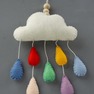 May include: A hanging mobile featuring a white felt cloud with two small wooden beads at the top. Seven felt raindrops in red, yellow, green, light blue, dark blue, purple, and cream hang from the cloud by white strings at varying lengths.