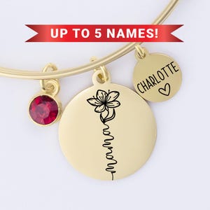 May include: Gold bangle bracelet with charms. The charms include a red gemstone, a flower charm, and a charm with the name "CHARLOTTE" engraved on it. The bracelet has a sign that says "UP TO 5 NAMES!"