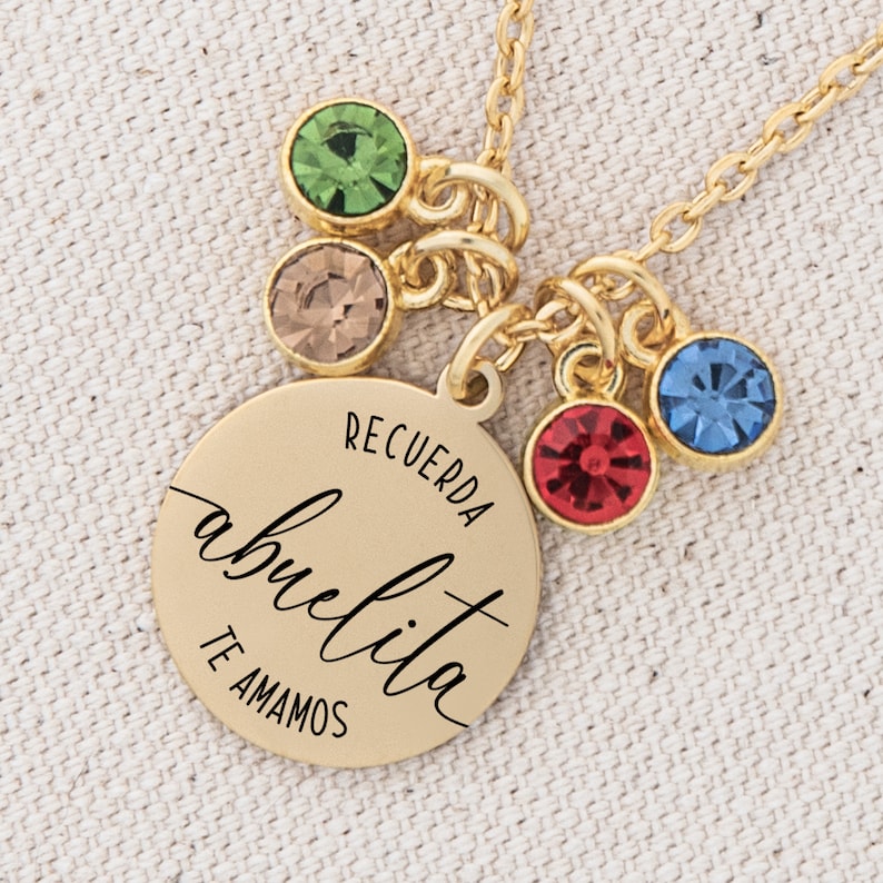 May include: Gold necklace with a round pendant that says "Recuerda abuelita te amamos". The pendant has a green, brown, red, and blue crystal charm.