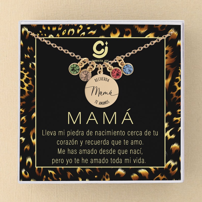 May include: A gold necklace with a round charm that says "Recuerda Mam&aacute; Te Amamos" in Spanish. The necklace has five colorful crystal charms. The necklace is in a white box with a leopard print background.