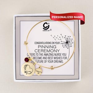 May include: A gold-colored bangle bracelet with a round charm featuring a stethoscope and floral design, and a smaller charm with the name "Charlotte RN". The bracelet is presented in a white box with text that reads "Congratulations on your Pinning Ceremony".