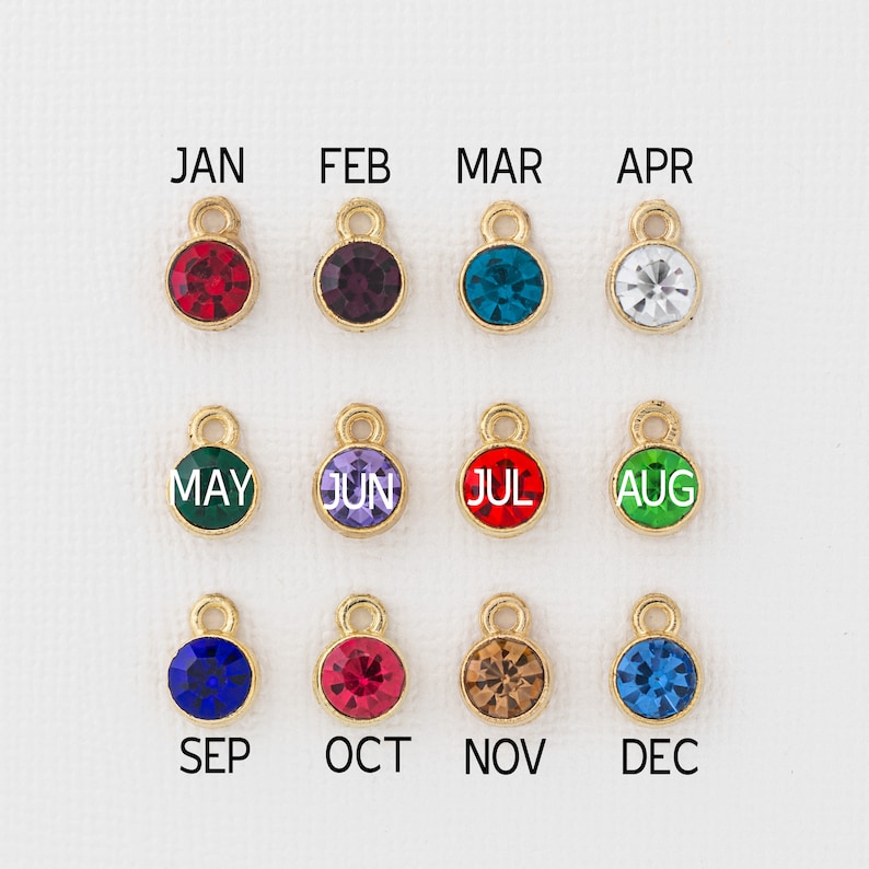 May include: Twelve gold charms with different colored crystals and the months of the year written on them.  The charms are arranged in three rows of four.  The crystals are red, purple, blue, clear, green, lavender, red, green, blue, red, brown, and blue.