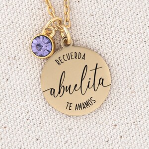 May include: Gold necklace with a round pendant that says "Recuerda abuelita te amamos" in black lettering. The pendant has a small purple gemstone attached to the top.