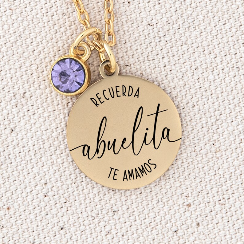 May include: Gold necklace with a round pendant that says "Recuerda abuelita te amamos" in black lettering. The pendant has a small purple gemstone attached to the top.
