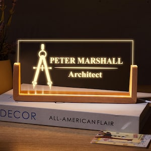 Könnte beinhalten: Ein personalisiertes, beleuchtetes Schreibtischschild mit dem Namen "PETER MARSHALL" und dem Titel "Architekt" in weißer Schrift. Das Schild zeigt ein leuchtendes Kompassdesign und steht auf einem Holzsockel.