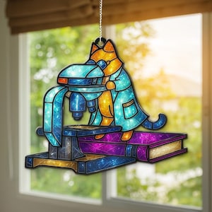 May include: A stained-glass style ornament featuring a cat in a lab coat using a microscope. The cat is orange and yellow, the microscope is blue, and the base is purple and gold. The ornament hangs from a silver chain.
