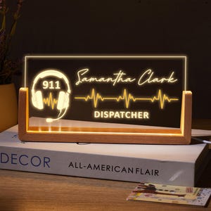 May include: A personalized LED desk lamp with a wooden base. The lamp features the name "Samantha Clark", the number "911", a headset graphic, and the word "DISPATCHER" in a glowing, warm white light. The lamp sits on a book.