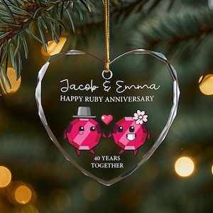 Pode incluir: Enfeite de vidro transparente em forma de coração com o texto "Jacob & Emma" e "Happy Ruby Anniversary". Dois personagens de rubi de desenho animado de mãos dadas, com o texto "40 Years Together" abaixo. O enfeite pende de um fio dourado.