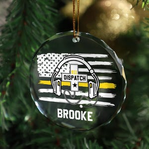 May include: A clear, round Christmas ornament with a gold string. The ornament features a distressed American flag design with a yellow stripe, headphones, and the text "911 DISPATCH" and "BROOKE".