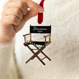May include: A brown wooden director's chair with a black seat and back. The chair is decorated with colourful string lights and a black sign that says "Steven 2024".