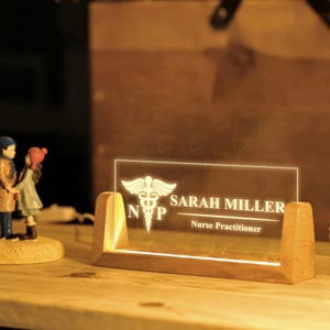 May include: A lighted desk nameplate with a wooden base. The clear acrylic panel displays the name "SARAH MILLER" and "Nurse Practitioner" below a medical symbol. The nameplate is illuminated with a warm glow.