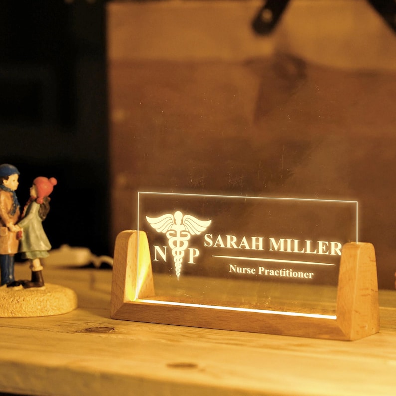May include: A lighted desk nameplate with a wooden base. The clear acrylic panel displays the name "SARAH MILLER" and "Nurse Practitioner" below a medical symbol. The nameplate is illuminated with a warm glow.