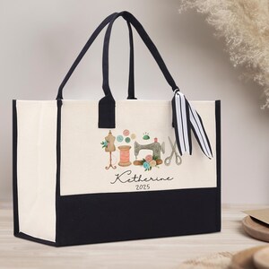 Peut inclure: Sac fourre-tout cr&egrave;me avec un fond et des poign&eacute;es noirs. Il pr&eacute;sente une illustration &agrave; l'aquarelle d'outils de couture, dont une machine &agrave; coudre, des ciseaux et des bobines de fil. Le nom "Katherine" et l'ann&eacute;e "2025" sont &eacute;crits en dessous.