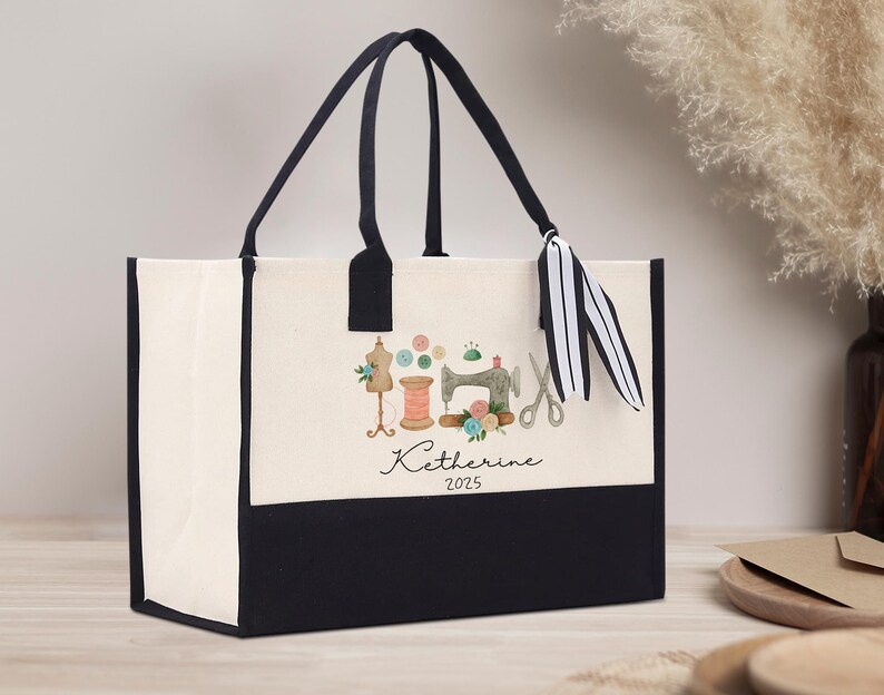 Peut inclure: Sac fourre-tout cr&egrave;me avec un fond et des poign&eacute;es noirs. Il pr&eacute;sente une illustration &agrave; l'aquarelle d'outils de couture, dont une machine &agrave; coudre, des ciseaux et des bobines de fil. Le nom "Katherine" et l'ann&eacute;e "2025" sont &eacute;crits en dessous.