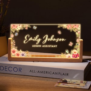 Puede incluir: Un letrero de escritorio de madera con un inserto de acrílico transparente que presenta un diseño floral con mariposas y el texto "Emily Johnson, Admin Assistant". El letrero está iluminado desde atrás, creando un brillo cálido.