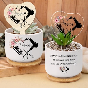Könnte beinhalten: Zwei weiße Keramik-Pflanzgefäße mit Holzuntersetzern, die jeweils eine Sukkulentenpflanze enthalten. Eines hat einen herzförmigen Holzpflock mit floralem Design, dem Namen "Jessica" und Salonwerkzeugen. Das andere hat einen transparenten herzförmigen Pflock mit dem gleichen Design und dem Text "Never underestimate the difference you make and the lives you touch."