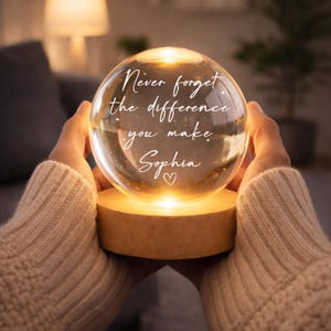 Puede incluir: Una esfera de cristal brillante con la inscripción "Never forget the difference you make Sophia" y un símbolo de corazón. La esfera se asienta sobre una base de madera. La esfera es sostenida por una persona que lleva un suéter beige. La esfera está iluminada desde dentro.
