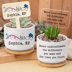 Może przedstawiać: Dwie białe ceramiczne doniczki z sukulentami, każda na drewnianej podstawce. Jedna doniczka ma drewniany znak z napisem "Sophia, NP" i ikonami medycznymi. Druga doniczka ma przezroczysty znak z cytatem.