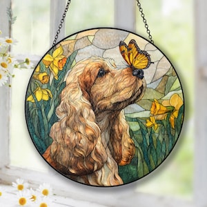 Puede incluir: Arte de ventana de vidriera redonda que representa un Cocker Spaniel con una mariposa en la nariz. El perro es de color marrón claro con pelo largo y ondulado. Los narcisos amarillos y el follaje verde rodean al perro. El arte de la ventana está suspendido por una cadena negra.