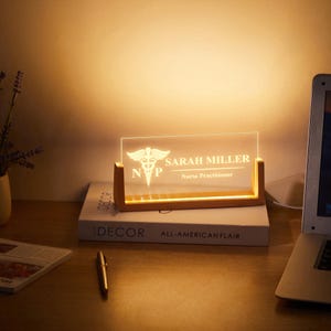 May include: A lighted desk nameplate with a wooden base. The clear acrylic panel displays the name "SARAH MILLER" and "Nurse Practitioner" with a medical symbol. The nameplate sits on a book with the word "DECOR" visible.