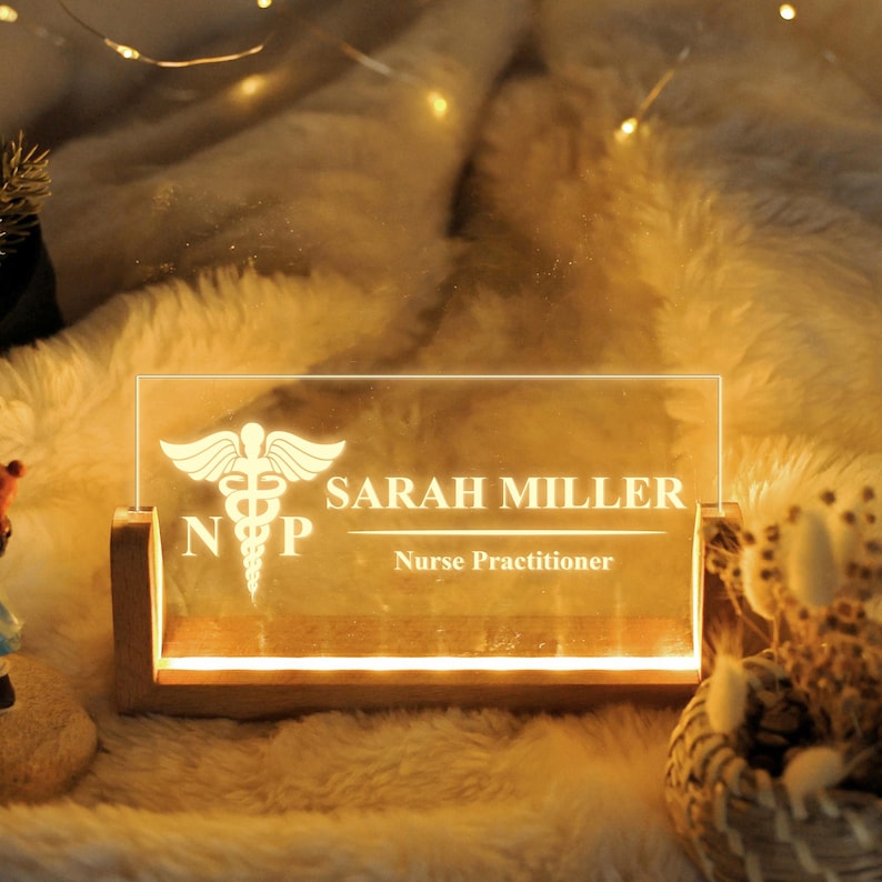 May include: A personalized LED desk sign with the name "SARAH MILLER" and the title "Nurse Practitioner" etched into a clear acrylic panel. The sign features the medical caduceus symbol and is set in a wooden base, illuminated with a warm glow.