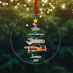 Peut inclure: Une décoration de Noël ronde et transparente avec un ruban rouge. L'ornement présente un motif de sapin de Noël composé d'outils, décoré de lumières colorées. Le nom "James" et l'année "2024" sont imprimés en bas.