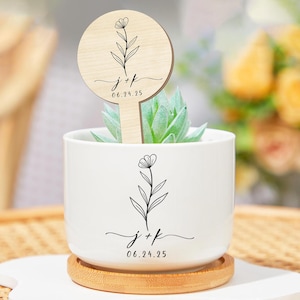 Puede incluir: Una maceta de cerámica blanca con una planta suculenta, sobre un posavasos de madera. La maceta y un marcador de plantas de madera presentan un diseño floral con las iniciales "j+k" y la fecha "06.24.25".