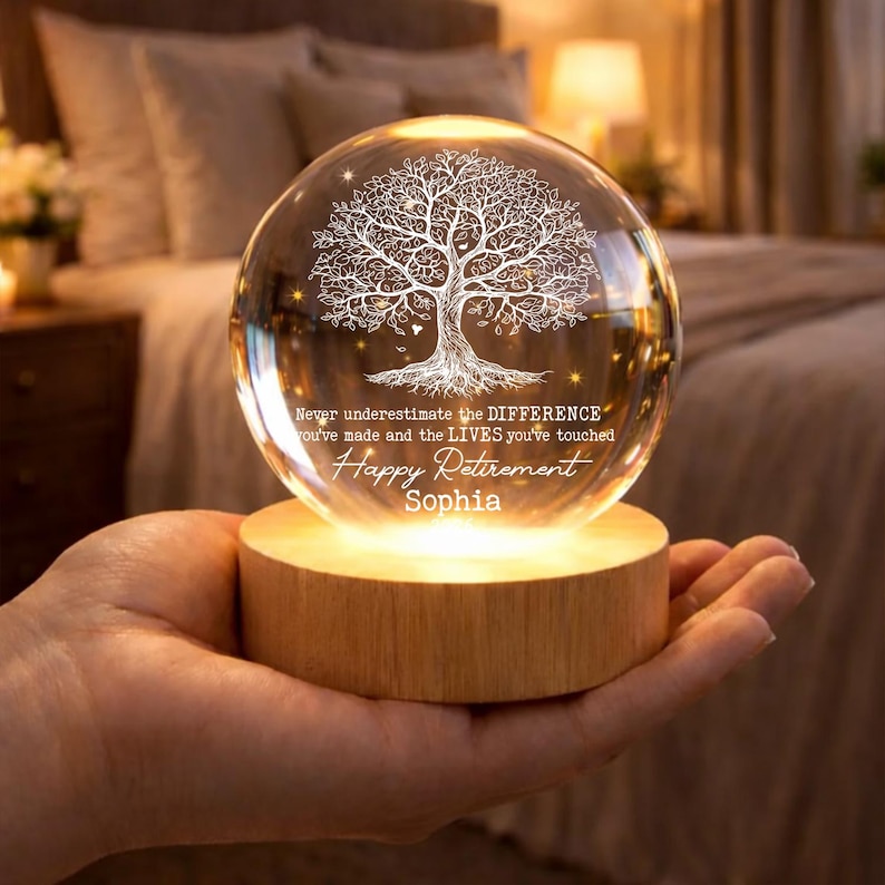 K&ouml;nnte beinhalten: Eine klare Glaskugel mit einem wei&szlig;en Baumdesign, die auf einem Holzsockel ruht. Die Kugel tr&auml;gt den Text "Never underestimate the DIFFERENCE you've made and the LIVES you've touched. Happy Retirement Sophia."