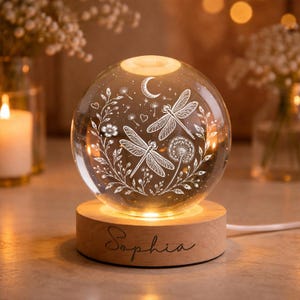 May include: A clear glass sphere lamp featuring an etched design of two dragonflies, a crescent moon, dandelion seeds, and floral vines. The sphere rests on a round, light-coloured wooden base with the name Sophia engraved in black script.