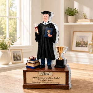 Op de afbeelding: Een afstudeerfiguur met een afgestudeerde in een zwarte toga en hoed, met een diploma en een blauw boek. De basis bevat de tekst "David", "He believed he could, so he did!", "Bachelor of Science" en "University of Michigan - Class of 2026". Er is ook een gouden trofee aanwezig.