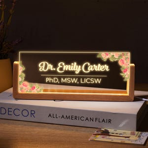 May include: A wooden desk sign with a clear acrylic insert. The insert features a floral design and the text "Dr. Emily Carter, PhD, MSW, LICSW".
