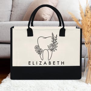 Peut inclure: Un sac fourre-tout en toile blanche avec des anses noires et un fond noir. Le sac présente un dessin au trait noir et blanc d'une dent avec des fleurs, et le texte "ELIZABETH" est imprimé en noir sous la dent.