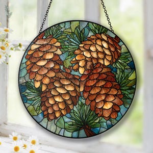 May include: Round stained glass panel featuring three detailed pine cones in shades of brown and gold, set against a backdrop of green and blue foliage. The panel is framed in black and hangs from a chain.