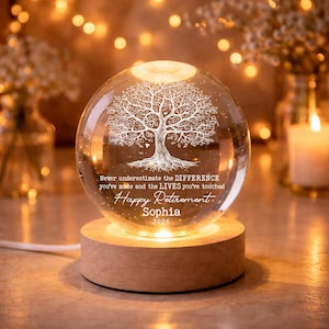 K&ouml;nnte beinhalten: Eine transparente Glaskugel mit einem detaillierten Baumdesign, von unten beleuchtet. Sie steht auf einem Holzsockel und tr&auml;gt den Text "Happy Retirement" und einen Namen, ideal als Geschenk zur Rente.
