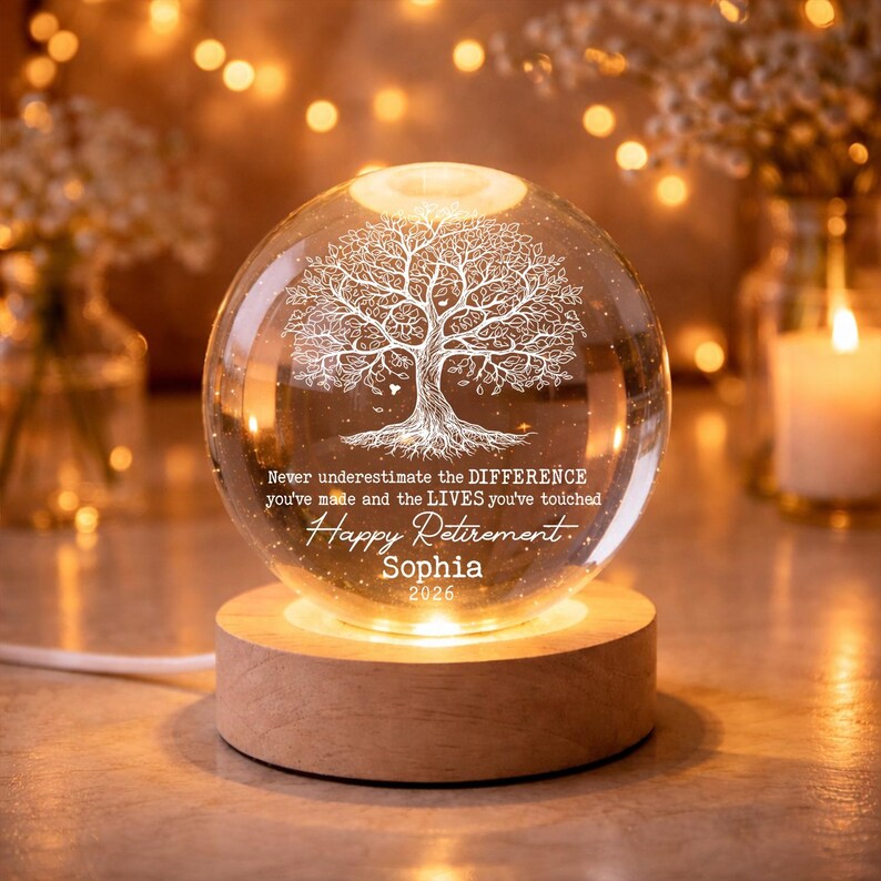 K&ouml;nnte beinhalten: Eine transparente Glaskugel mit einem detaillierten Baumdesign, von unten beleuchtet. Sie steht auf einem Holzsockel und tr&auml;gt den Text "Happy Retirement" und einen Namen, ideal als Geschenk zur Rente.
