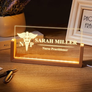May include: A lighted desk nameplate with a wooden base. The clear acrylic panel displays the name "SARAH MILLER" and "Nurse Practitioner" in a white font. A medical symbol and the letters "NP" are also visible.